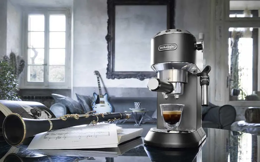 A compact De’Longhi espresso machine sits on a glossy table beside sheet music and a clarinet, with a guitar in the background.