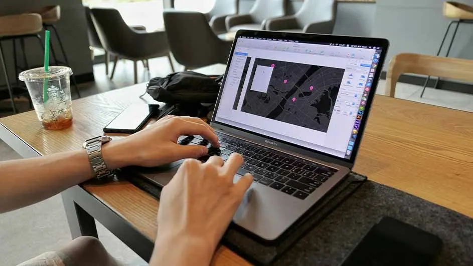 Person types on a laptop showing a map with pink markers. A drink, phone, and bag are on the wooden table. Chairs in background.