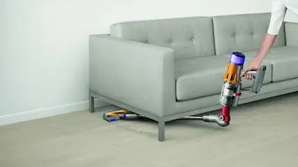 Een grijze bank staat op een houten vloer terwijl een persoon met een draadloze stofzuiger eronder schoonmaakt.