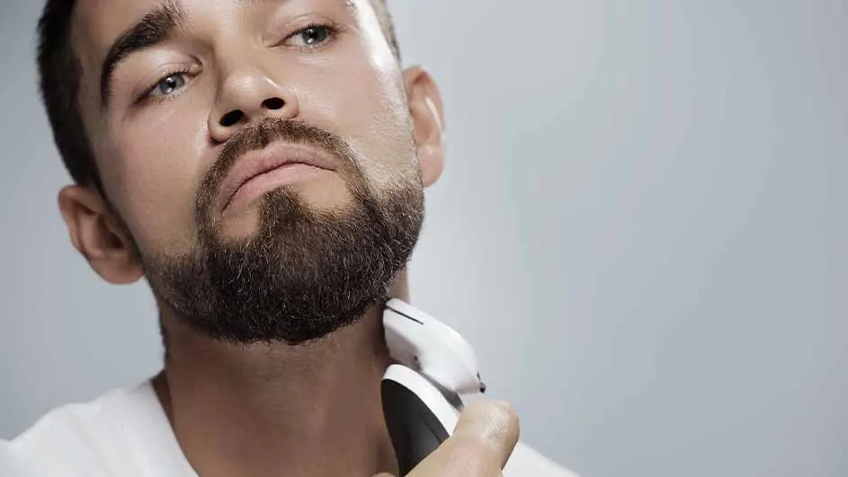 Een man trimt zijn baard met een wit en zwart elektrisch scheerapparaat tegen een grijze achtergrond.