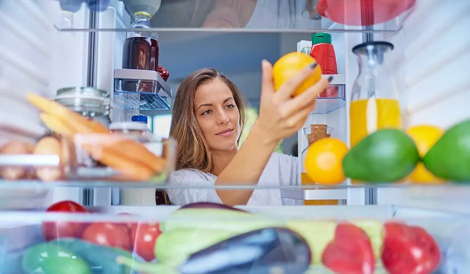 Een persoon pakt een citroen uit een goed gevulde koelkast met groenten, fruit, wortels, avocado’s, paprika’s en een fles sinaasappelsap.