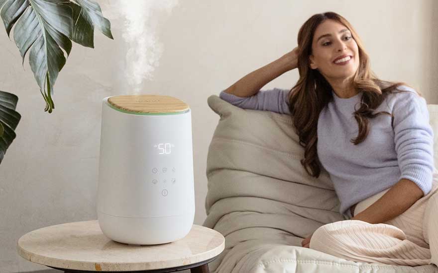 Een vrouw zit ontspannen op een bank, glimlachend naar een moderne luchtbevochtiger op een tafel die stoom verspreidt in de kamer.