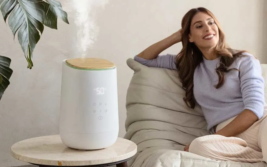Een vrouw zit ontspannen op een bank, glimlachend naar een moderne luchtbevochtiger op een tafel die stoom verspreidt in de kamer.