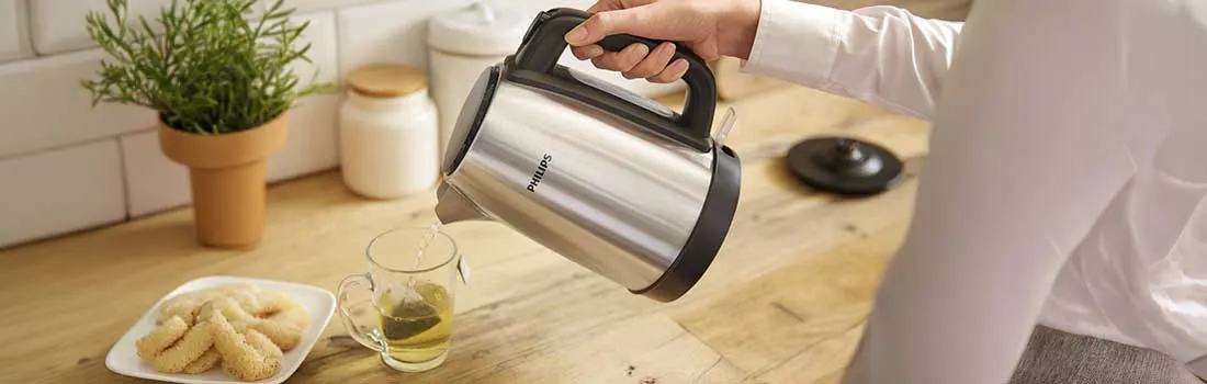 Hand schenkt heet water uit een roestvrijstalen Philips-waterkoker in een glazen kop thee, naast koekjes op een houten keukenblad.