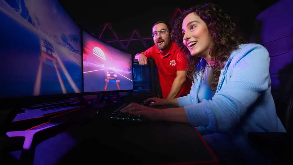 Een vrouw in een blauw vest speelt een racegame op een computer met meerdere monitoren. Een man in een rood shirt kijkt toe.