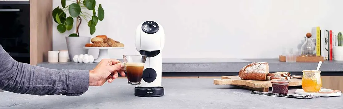 Een hand pakt een glas koffie bij een witte koffiemachine op een modern aanrecht, met brood, jam, honing en planten op de achtergrond.
