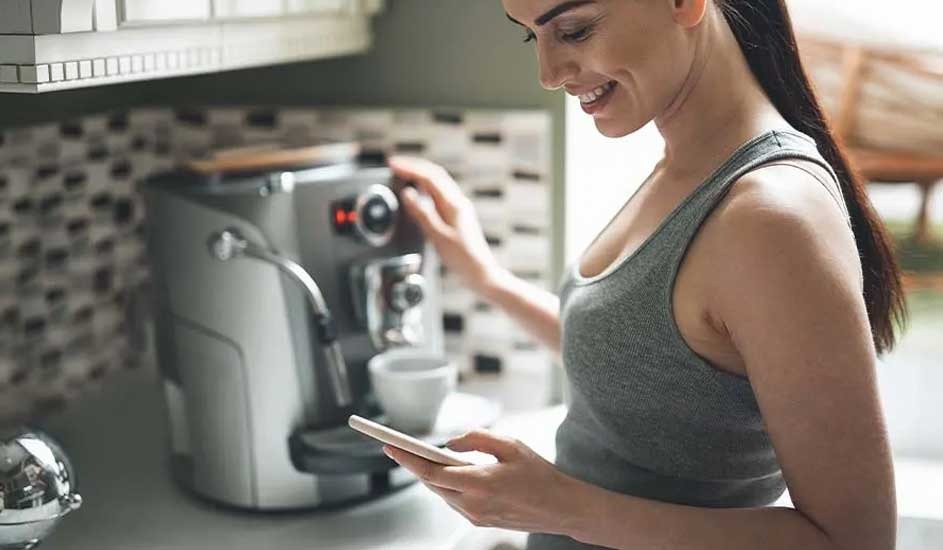 Woman uses a phone near a coffee machine. A white cup is under the machine. Kitchen counter and cabinets are visible.