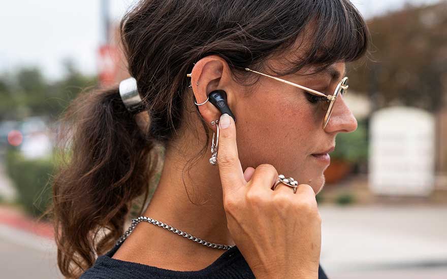 Side view of a person with a ponytail touching wireless earbuds, wearing glasses and jewelry.