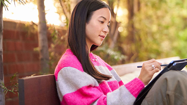 Een vrouw in een roze-wit gestreepte trui zit buiten op een bank en tekent op een tablet met een stylus.