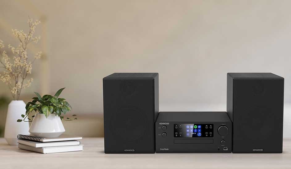 A black Kenwood stereo with two speakers sits on a table, next to stacked books, a plant and a white vase with branches.