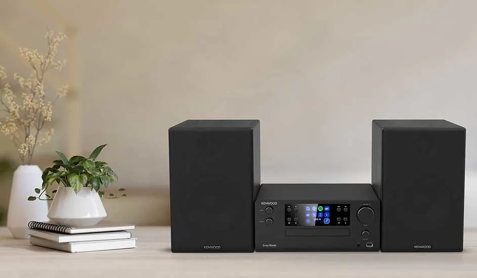 Een zwarte Kenwood stereo met twee speakers staat op een tafel naast boeken, een plantje en een witte vaas met takken.