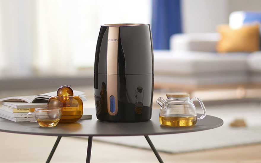 Een zwarte Philips luchtbevochtiger staat op een salontafel, naast glazen en een theepot, in een modern ingerichte woonkamer.