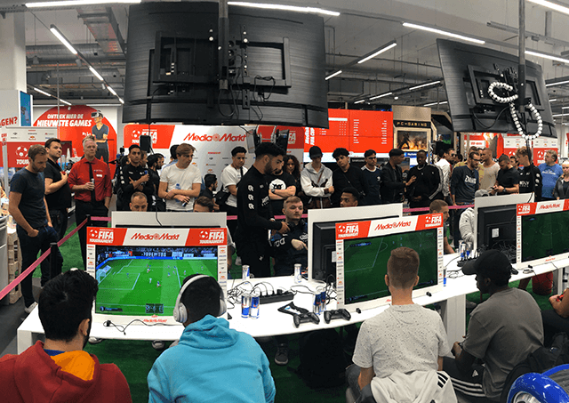 Mensen spelen FIFA op computers in een winkel met rode en witte reclame en hangende tv's.