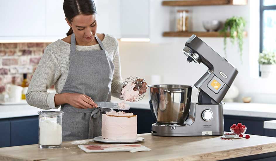 Een vrouw met grijs schort strijkt roze crème op een taart, naast een metalen keukenmixer, pot met bloem en frambozen op het aanrecht.