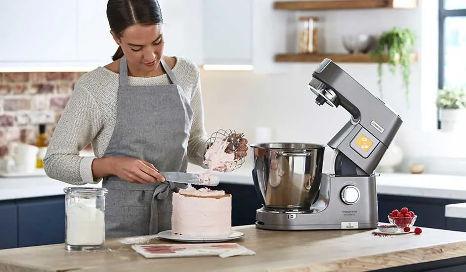 Een vrouw met grijs schort strijkt roze crème op een taart, naast een metalen keukenmixer, pot met bloem en frambozen op het aanrecht.