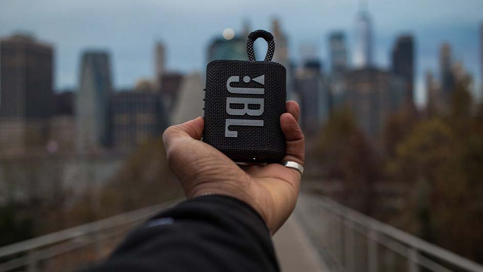 Een hand houdt een zwarte JBL-luidspreker vast met een lus, tegen een achtergrond van gebouwen en bomen.
