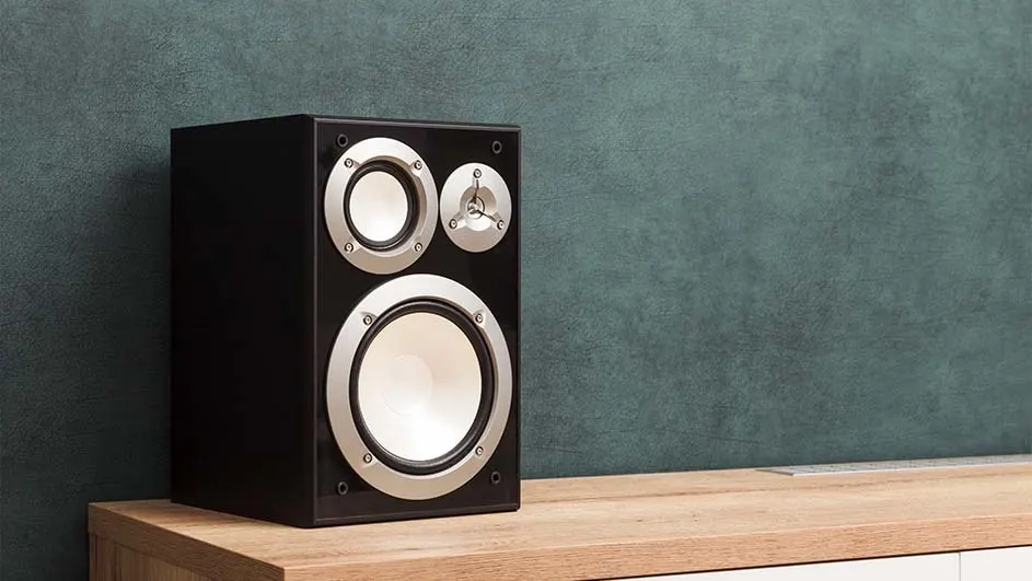 A black speaker with silver trim sits on a light wood surface against a textured teal wall.