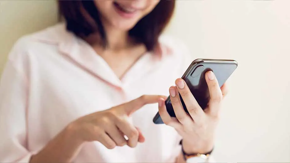 Vrouw in roze blouse gebruikt een zwarte smartphone. Ze draagt een horloge. De achtergrond is een effen muur.