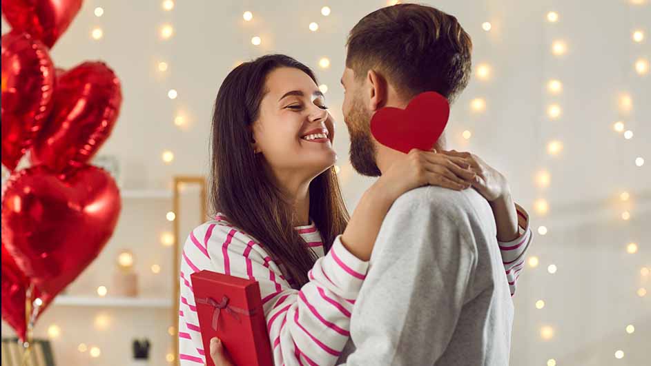 Een vrouw en man omhelzen elkaar, met rode hartjesballonnen en cadeautje, tegen een achtergrond van feestverlichting.