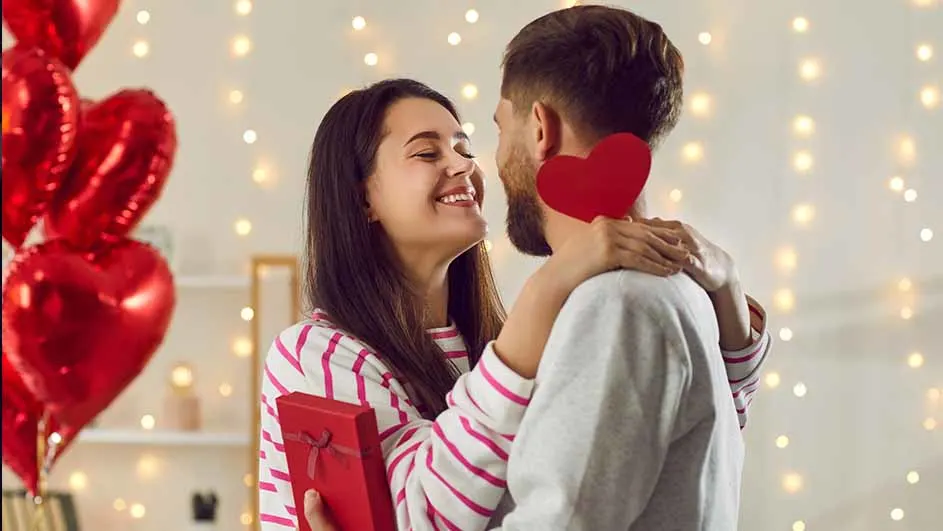 Een vrouw en man omhelzen elkaar, met rode hartjesballonnen en cadeautje, tegen een achtergrond van feestverlichting.