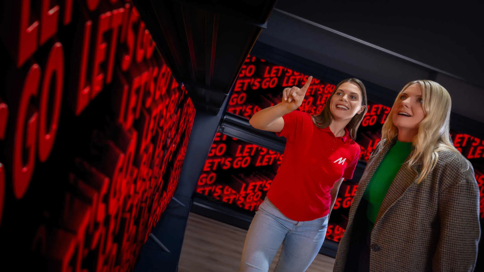Twee vrouwen staan voor een groot scherm met rode tekst “LET’S GO”; één vrouw draagt een rood shirt en wijst naar het scherm.