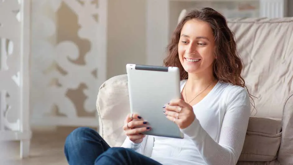 Vrouw zit in stoel met een tablet in haar handen. Op de achtergrond een witte muur met decoratie.