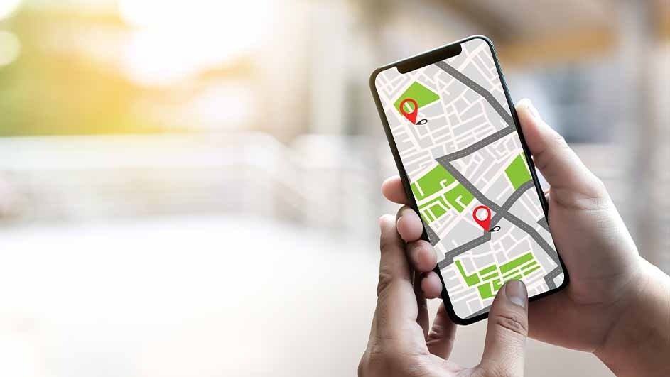 A person holds a smartphone displaying a map with green areas and gray roads marked by two red location pins.
