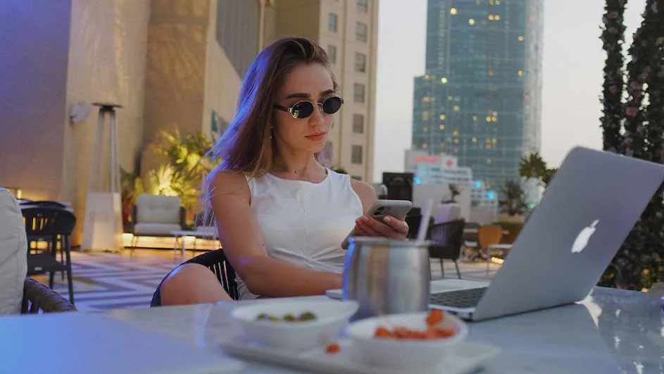 Een vrouw in een witte top en zonnebril zit aan een tafel met een laptop en telefoon, met een stadsgezicht op de achtergrond.