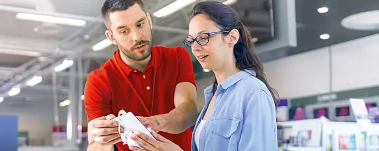 Een man in een rood shirt laat een vrouw met een blauw shirt een witte telefoon zien in een winkel.