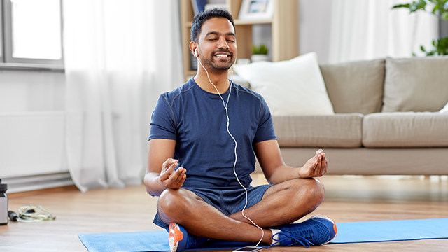 Een man zit gekruist op een blauwe mat met een blauw shirt en oortjes, in een lichte kamer met een bank en een raam.