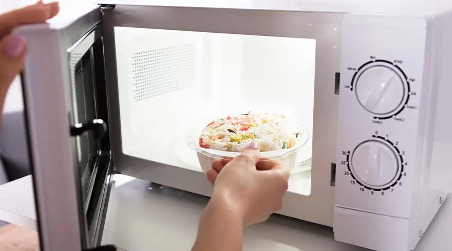 Een openstaande witte magnetron met twee draaiknoppen. Een hand plaatst een transparante kom met rijst in de magnetron.