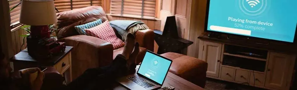 Woonkamer met laptop op een tafel, voeten omhoog, tv met wifi-symbool en zachte bruine stoelen.