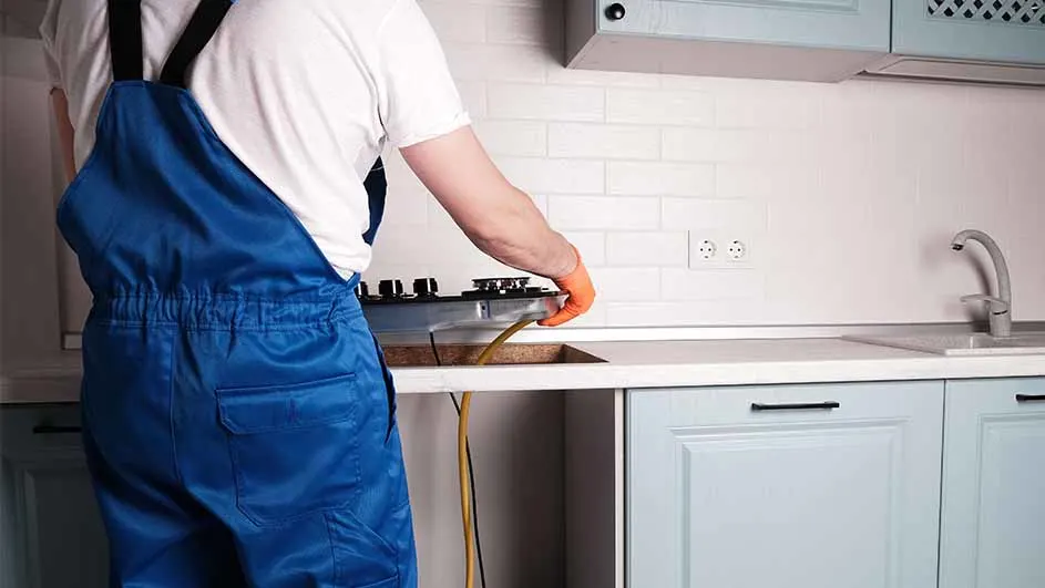 Een monteur in blauwe overall installeert een kookplaat in een lichtblauwe keuken met witte tegels.