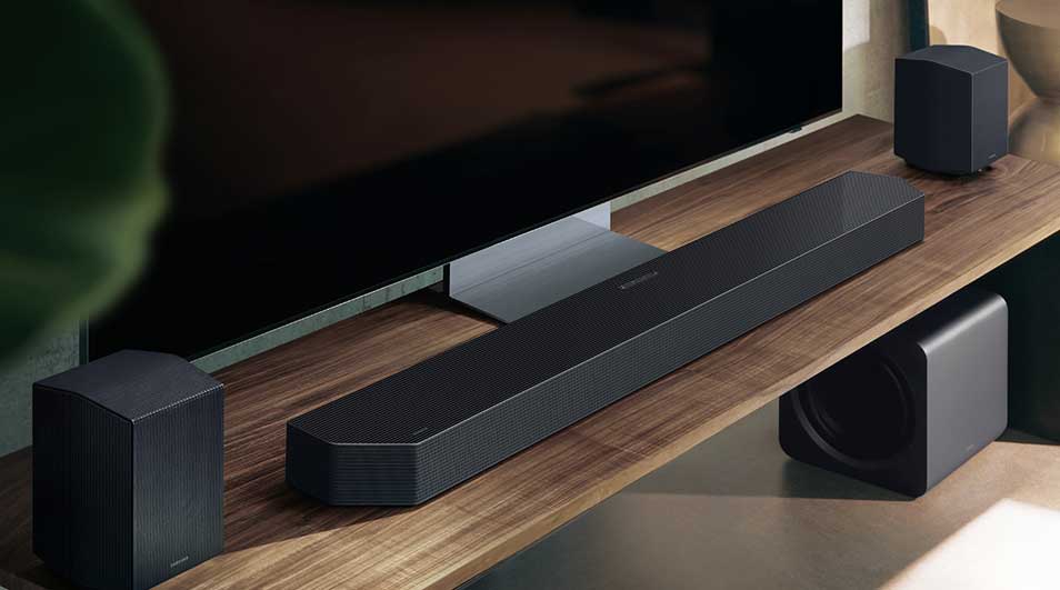 Op een houten tv-meubel staan een soundbar, twee achterspeakers en een subwoofer naast een grote flatscreen tv, allemaal in zwart design.