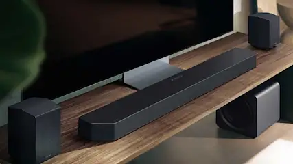 Op een houten tv-meubel staan een soundbar, twee achterspeakers en een subwoofer naast een grote flatscreen tv, allemaal in zwart design.