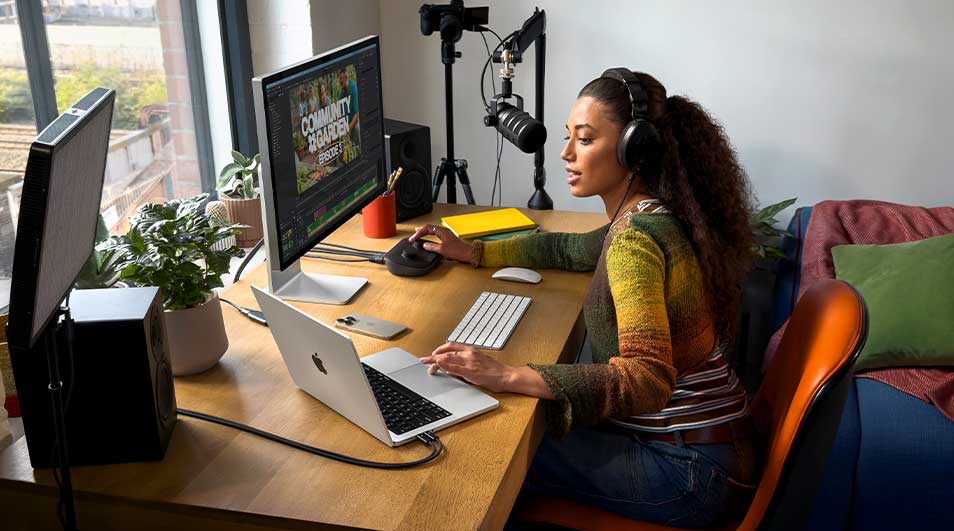 Woman wearing headphones creates content at a desk, using a laptop, large monitor, microphone and speakers in a bright studio corner.
