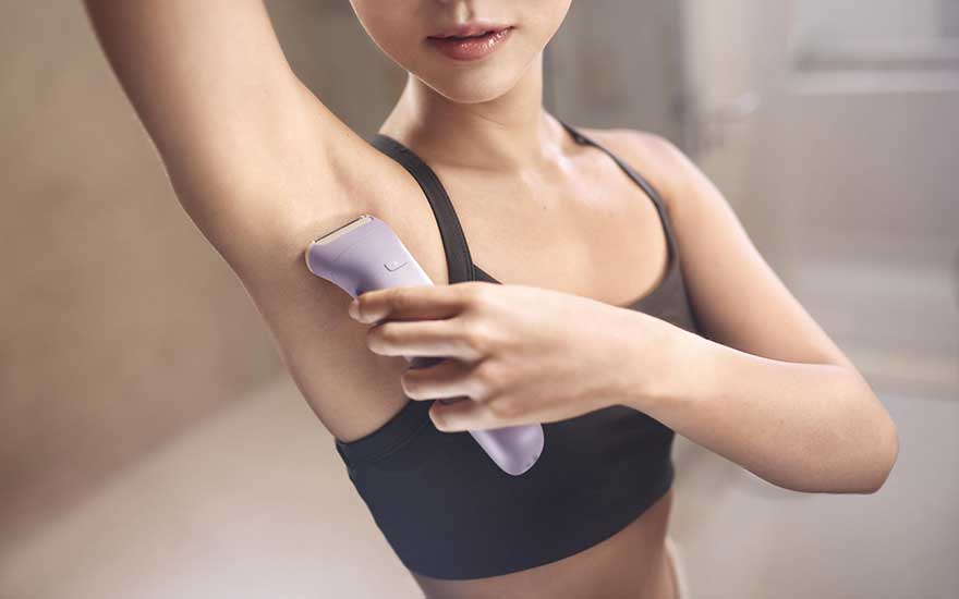 A young woman in sportswear shaves her armpit with a lilac electric shaver in a bright bathroom setting.
