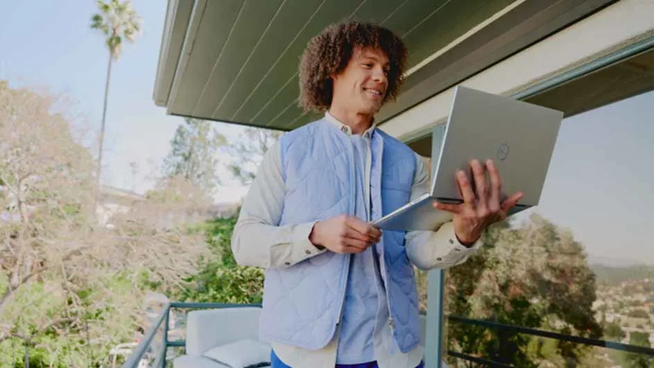 Man glimlacht en houdt een open laptop vast op een balkon. Op de achtergrond staan bomen en een palmboom.