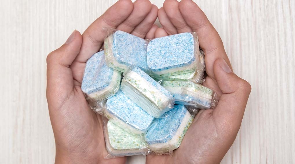 Two hands hold blue and green dishwasher tablets, individually wrapped in plastic, against a white wood-patterned background.