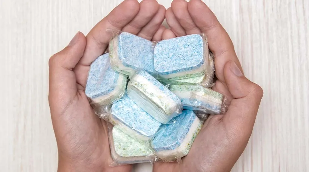 Two hands hold blue and green dishwasher tablets, individually wrapped in plastic, against a white wood-patterned background.