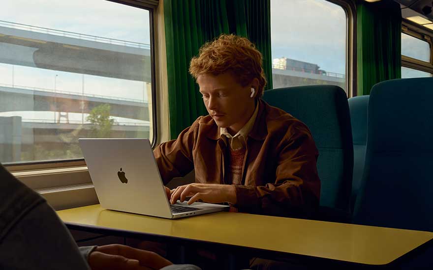 A man is working on his laptop on a train, wearing wireless earbuds. Through the window, a motorway and buildings are visible.