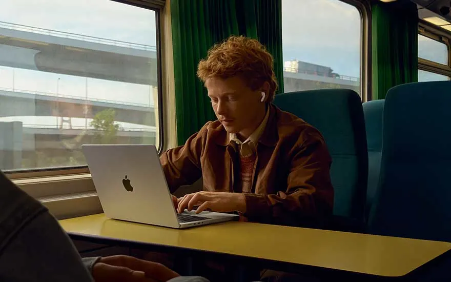 A man is working on his laptop on a train, wearing wireless earbuds. Through the window, a motorway and buildings are visible.