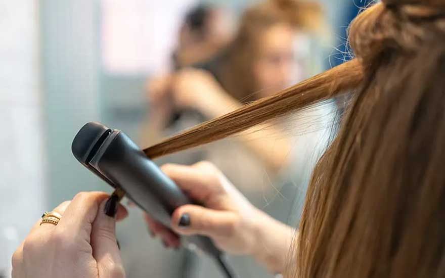 Close-up van een hand die een lok haar met een stijltang glad maakt, met een wazige salonachtergrond op de achtergrond.