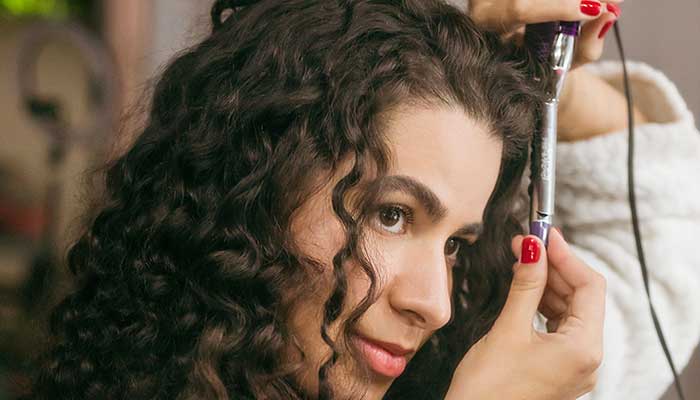 Vrouw met gekruld haar gebruikt een krultang met paarse accenten en rode nagellak voor haarstyling.