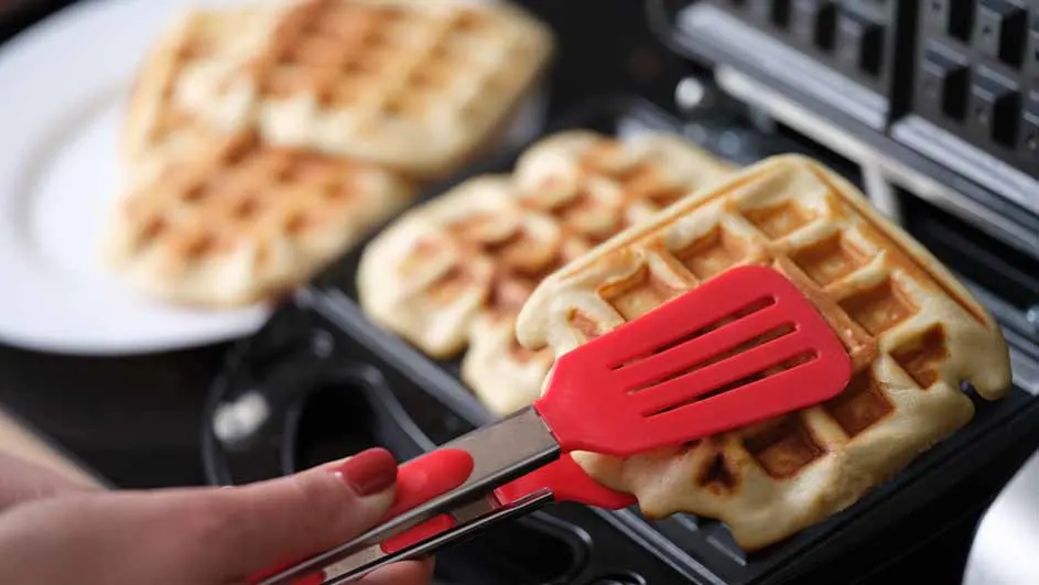 Een wafel wordt met een rode spatel uit een wafelijzer gehaald; andere wafels op een wit bord op de achtergrond.
