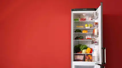 Open witte koelkast staat voor een rode achtergrond. Binnenin liggen groenten en fruit op glazen planken.