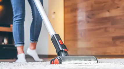 Een persoon stofzuigt een zacht tapijt met een moderne stofzuiger; alleen de benen zijn zichtbaar, de vloer oogt fris en netjes.