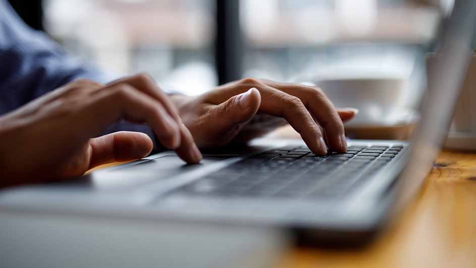 Twee handen typen op een laptop. De laptop staat op een houten tafel. Op de achtergrond is een kopje te zien.