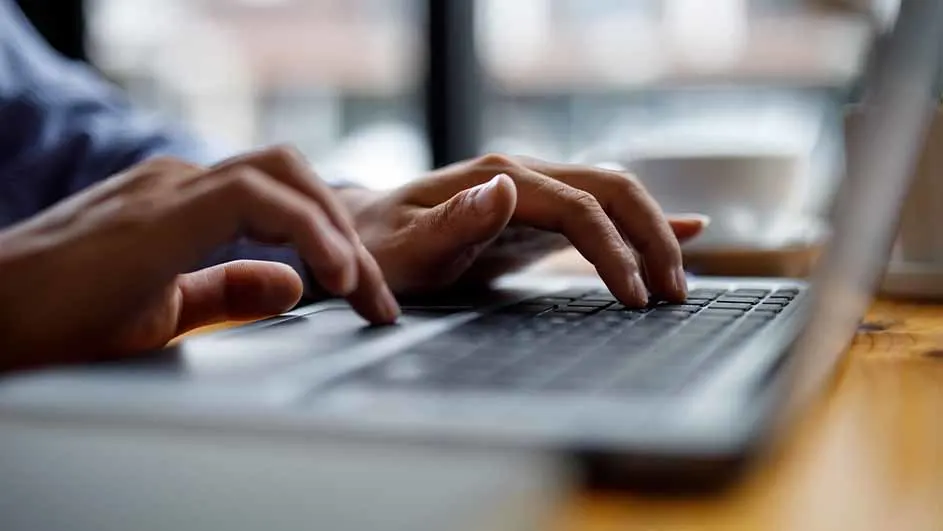Twee handen typen op een laptop. De laptop staat op een houten tafel. Op de achtergrond is een kopje te zien.