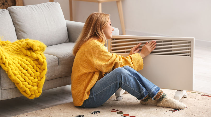 Vrouw zit op vloer, leunt tegen witte radiator. Ze draagt een gele trui en jeans. Op de bank ligt een geel deken.